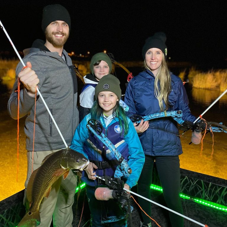 Family holding bows and a fish on a lit bowfishing boat at night