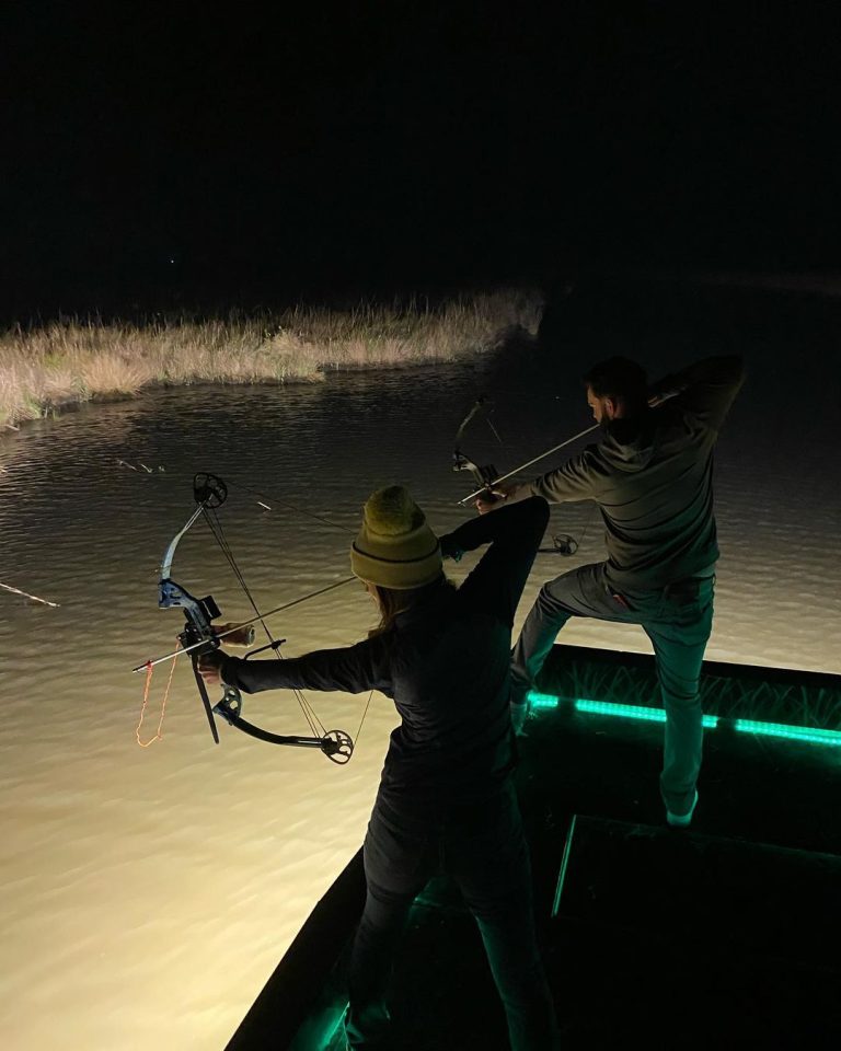 Two bowfishermen drawing their bows from a lit boat deck at night
