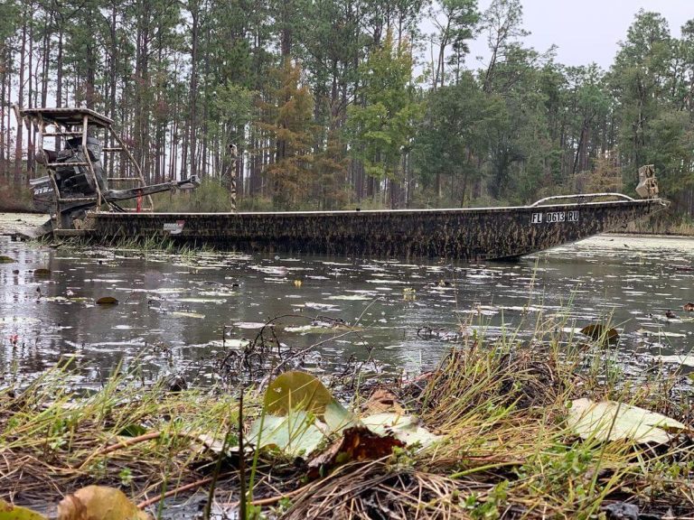 Camo mud boat with a raised platform on flooded water in a pine forest
